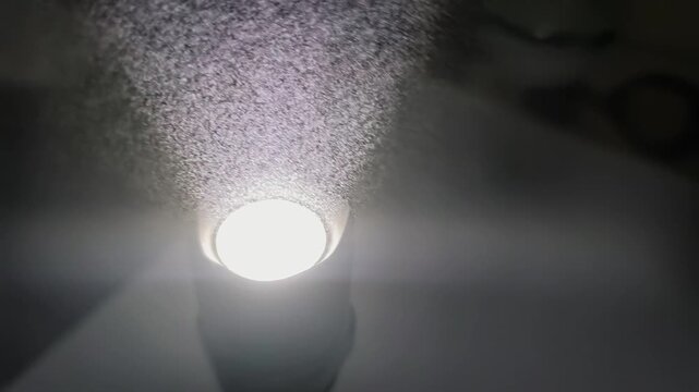 Bright flashlight beam cuts through darkness, revealing swirling dust particles suspended in the air. The intense cone of light highlights microscopic debris floating in dark space.
