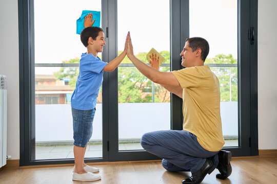 Happy mixed race father and son giving each other a high five while cleaning at home. Little hispanic boy helping his dad wash windows. Parent and child joining hands to celebrate achievement at home