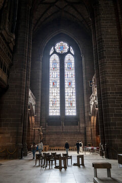 View of the interior of Liverpool Cathedral featuring massive stone arches, a towering stained glass window, and people walking across the floor in Liverpool, England, United Kingdom.