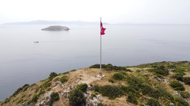 Aerial view of the Turkish flag waving proudly on a flagpole. The pole is on a scenic hill overlooking the Aegean Sea with islands in the distance