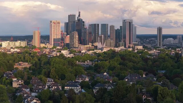 Frankfurt skyline glowing during golden hour sunset with modern skyscrapers and residential buildings surrounded by green trees, metropolitan urban landscape