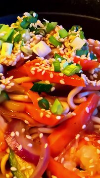 Extreme close up of a vibrant Asian noodle bowl packed with udon, red pepper, carrot and sesame seeds, capturing the color and texture of Asian street food