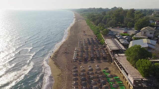 Drone flies over Kourouta beach, Greece. Sandy shoreline, beach cafe-bar umbrellas and bathers visible. Clear sunny summer day