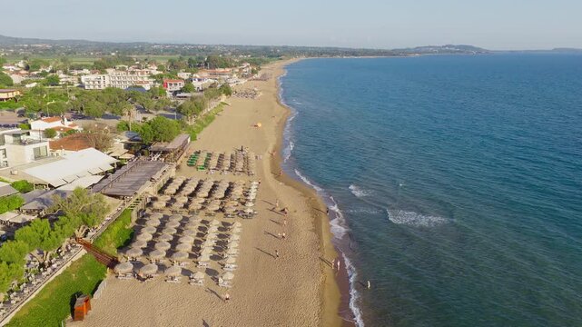 Aerial drone shot over Kourouta beach, Peloponnese, Greece. Beach cafe-bar umbrellas, bathers on sandy shoreline. Clear sunny summer day