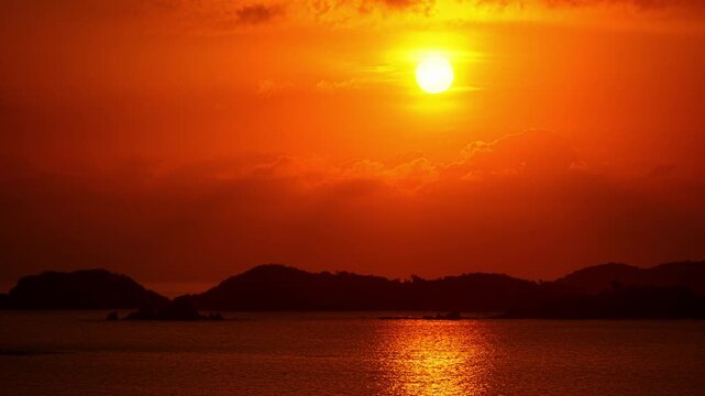 Orange sunset above coastal islands reflected across calm ocean waters at dusk as sun sets below clouds, silhouette of coastline, backdrop