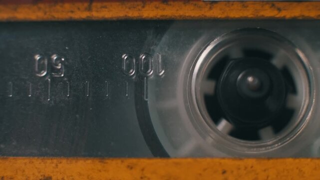 Extreme close-up macro of an audio cassette reel spinning inside an old yellow tape player. Worn plastic, counter numbers, and dust particles add to the nostalgic aesthetic.