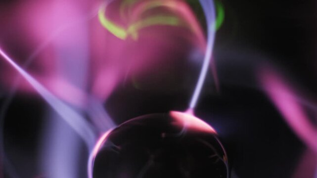 Extreme close-up of a central electrode emitting pink and purple plasma filaments. White electric arcs swirl through ionized gas streams with green light accents on a dark backdrop.