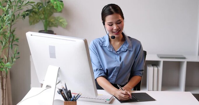 Friendly young asian woman working as a customer service representative, wearing a headset and smiling while providing support, assistance, and taking notes in a busy call center environment
