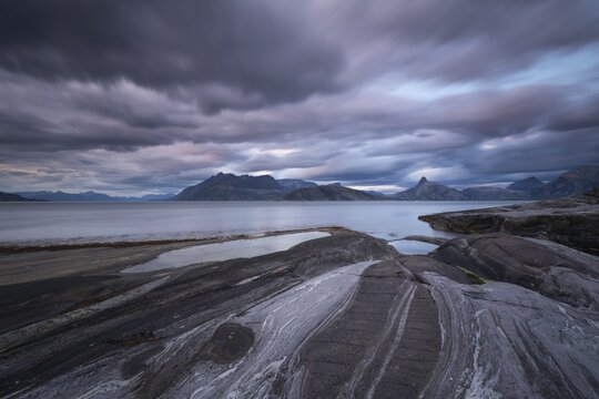 The Vestfjord in Nordland, Norway, offers a dramatic atmosphere near Bod&oslash;. Dark clouds are illuminated by the setting sun in shades of red and yellow. The rugged cliffs and mountains are reflected in the quiet fjord, which offers a breathtaking view of Lofoten
