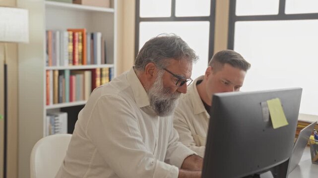 Man writing with pen at computer monitor while colleague leans over the desk, bookshelf and sunlight visible through window in building; collaboration focus.