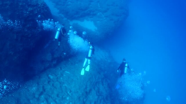  scuba divers exploring around a reef underwater deep blue water big rocks and bubbles ocean scenery 