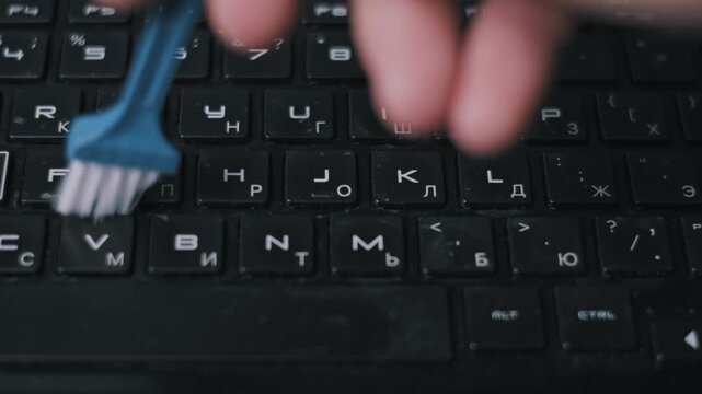 Top-down view of a hand sweeping dirt and crumbs from a dusty black computer keyboard with a small blue brush. The meticulous cleaning promotes hygiene and device longevity.