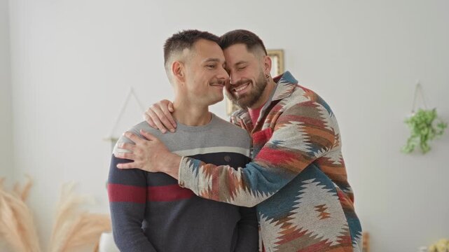 Two men embrace with arm around shoulder and cheek kiss in a studio with patterned sweater, earring and houseplants; affection intimacy.