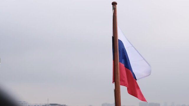 Russian flag flutters on a flagpole under a cloudy sky, displaying the blue, white, and red tricolor pattern in a serene outdoor setting