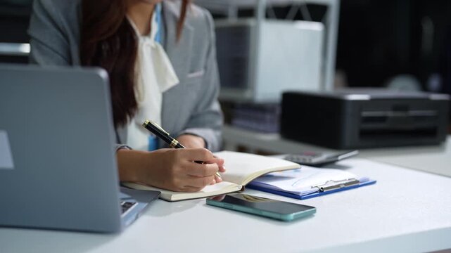 Business woman, taking notes sitting at desk using smartphone and laptop searching for information at work.