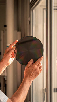 Hands inspecting dark circular silicon wafer disc against bright window in laboratory