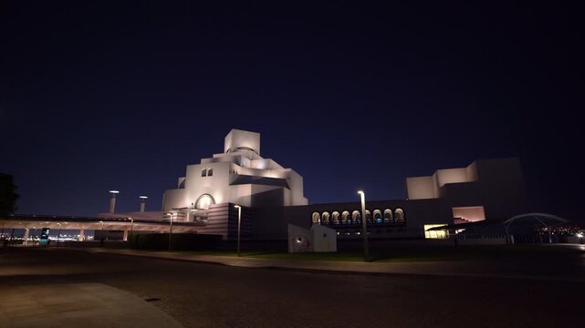 Wide night shot of the Museum of Islamic Art in Doha, Qatar, captured from street level, showcasing its striking geometric white facade softly illuminated against a deep blue night sky.
