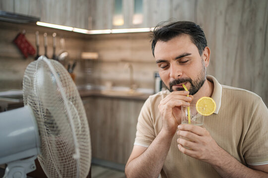 Man refreshing with a cold drink in front of a fan during a summer heat wave at home