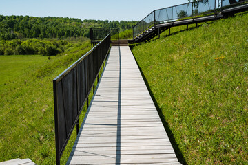 Fototapeta premium Serene outdoor walkway. Tranquil pathway with scenic views. Gentle hill trail shaded by calming summer sunlight. Calm journey along wooden route bordered by dark railing during summer