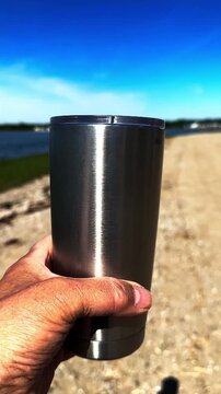 Close-up of a man's hand holding a reusable stainless steel travel mug on a sunny day. The background shows a gravel path near water