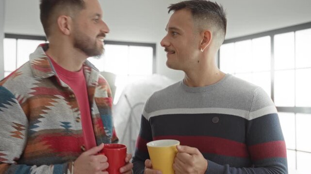 Two men touching noses and smiling while holding coffee mugs beside packed boxes in a building; affection moving day.