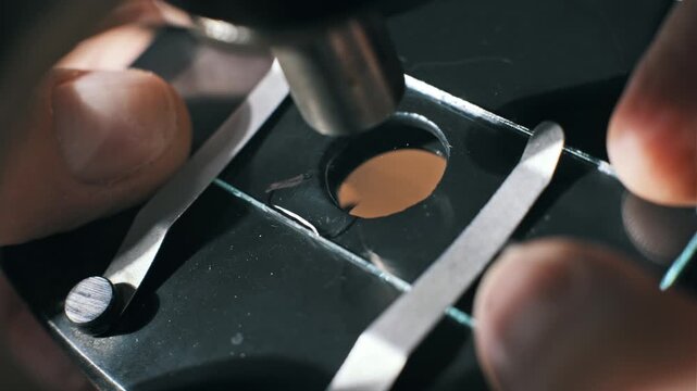 Top-down macro of fingers adjusting a glass slide with a brown liquid sample as a pipette adds clear fluid. The mixture creates a reaction for scientific analysis.