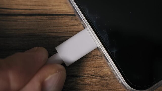 Extreme close-up of fingers inserting a white USB-C connector into a modern smartphone port. The device rests on a wooden table while recharging its battery with electric energy.