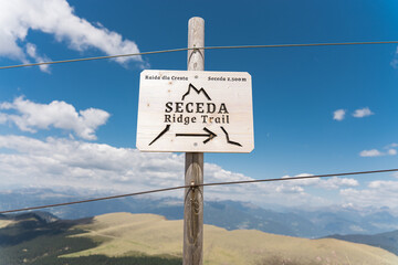 Naklejka premium Wooden directional sign for Seceda Ridge Trail on a mountain pass with panoramic alpine landscape view under blue sky, outdoor hiking and trekking navigation concept