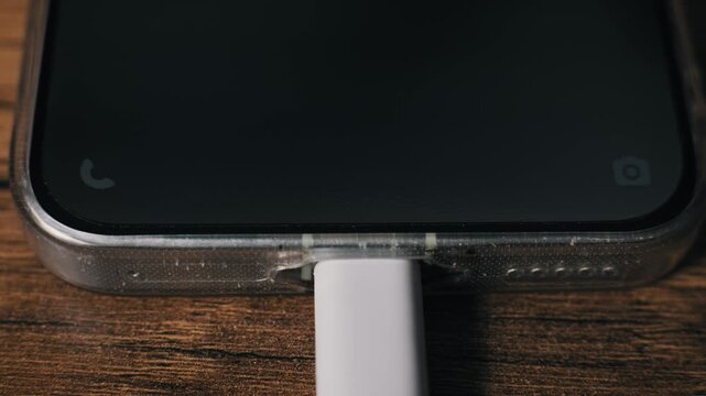 Close-up of a USB-C cable plugging into a smartphone charging port on a wood texture background. A blue electric arc animation appears on the screen indicating battery recharge.
