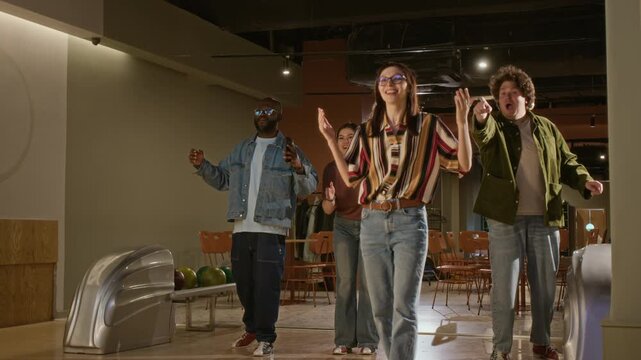 Caucasian woman throwing bowling ball while diverse young friends cheer and celebrate successful strike in bowling alley. Lifestyle advertisements, entertainment promos, and friendship campaigns