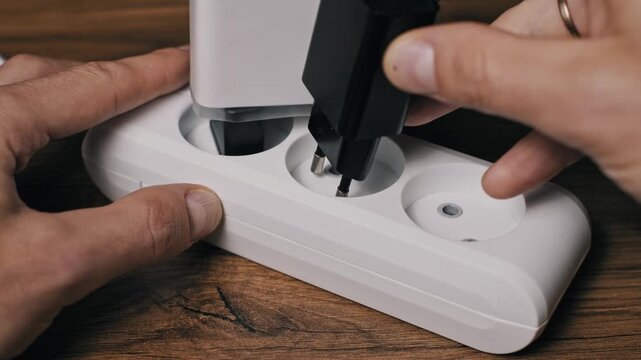 Hands plug multiple devices including a white charger and black grounded cord into a white triple socket power bar. The setup on a wood desk shows power consumption and usage.