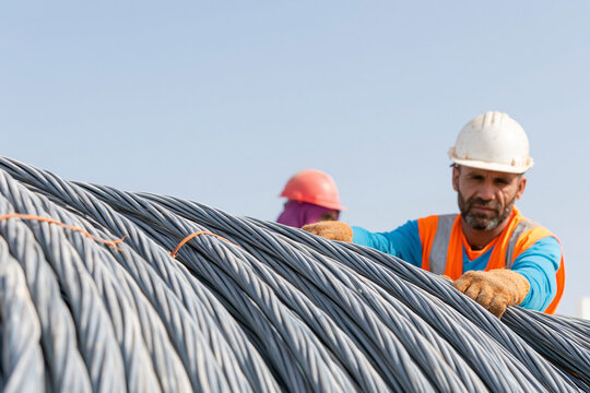 Construction worker in hard hat and safety vest handling steel wire coils at a construction site under clear blue sky with another worker in the background