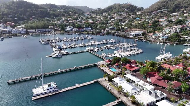 ow altitude drone approach over luxury sailing yachts and white catamarans moored at the docks of the sheltered Carenage marina in Saint George's town center, Grenada.
