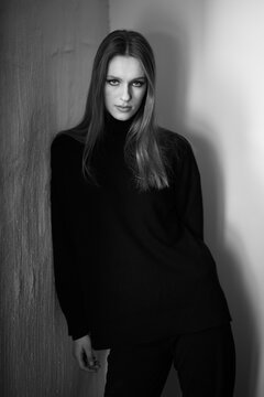 Dramatic black and white portrait of a woman leaning against a wall. The deep contrast, structured lighting, and strong gaze create a sophisticated and moody aesthetic