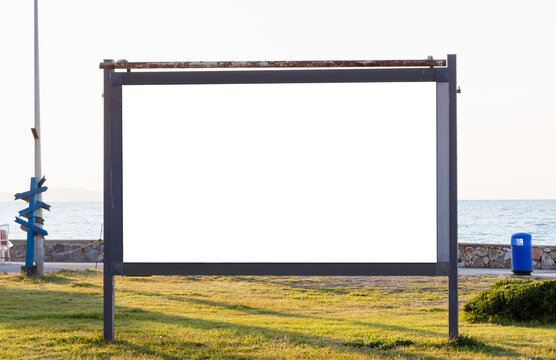 Large horizontal blank white outdoor advertising billboard mockup on a green grass field near a coastal promenade with sea view under a bright evening sky.