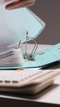 Vertical video hand inserting punched papers into a light blue ring binder, organizing important office documentation and business records on a desk with a calculator