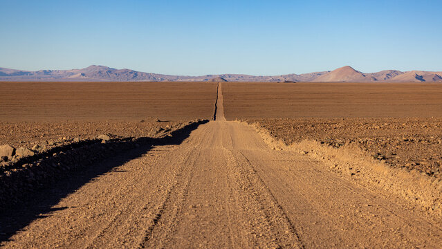 View of a long straight dirt road stretching through the vast arid landscape of the Atacama Desert under a clear blue sky San Pedro de Atacama, Antofagasta, Chile.