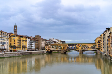 Fototapeta premium Iconic Ponte Vecchio bridge lined with shops spans the Arno River in Florence, Italy, with colorful buildings and a tower visible