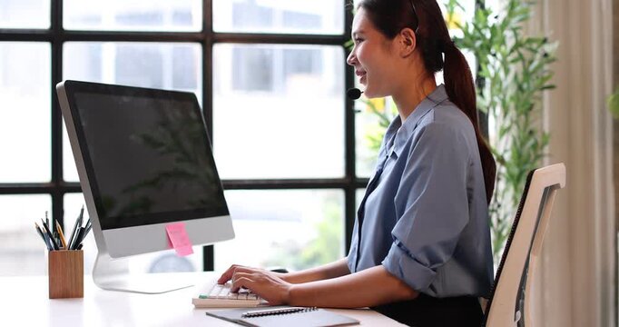 Friendly young asian woman working as a customer service representative, wearing a headset and smiling while providing support, assistance, and taking notes in a busy call center environment