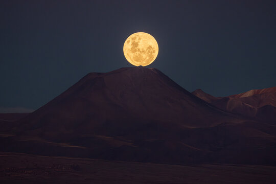 View of a glowing full moon rising directly above a volcanic mountain peak in the Atacama Desert under a twilight sky in San Pedro de Atacama, Antofagasta, Chile.