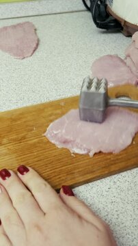 A close-up view shows female hands pounding a slice of raw meat with a metal tenderizer on a wooden cutting board. Concept of home cooking preparation, culinary skills, and making dinner.