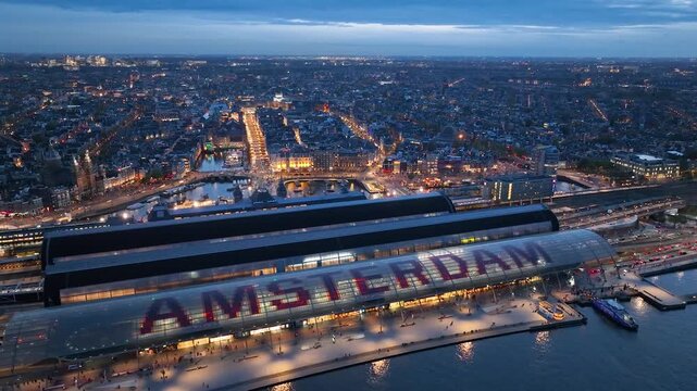 Aerial night drone panorama of Amsterdam, Netherlands. In the foreground is the central train station with Amsterdam sign on its roof. Narrow canals, bridges, streets, architectural lines of houses.4K