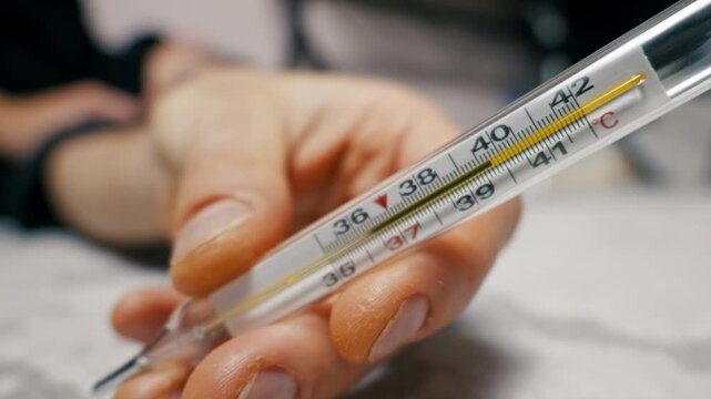 Hand holds a glass mercury thermometer showing a temperature of 39 degrees Celsius, indicating a fever. Camera pulls focus from a blurry background to the sharp thermometer in the foreground.