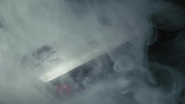 Retro audio cassette tape with a side 'A' label lies on a dark surface. Thick, white smoke billows and swirls across the object, slowly revealing it in a mysterious, moody light.