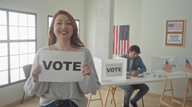 Woman holds vote sign with both hands while smiling, man at table fills ballot forms beside clear box and us flags inside a polling building; civic duty optimism.