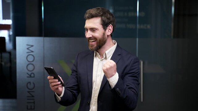 Excited businessman in suit standing in office reading great news on smartphone. Smiling manager celebrating success, feeling surprised and happy after receiving positive business message or result