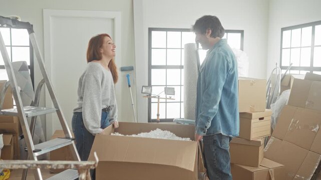 Woman throws packing peanuts at man while both smile and unpack a large cardboard box amid stacked moving boxes, ladder and bubblewrap inside a building; playful teamwork.