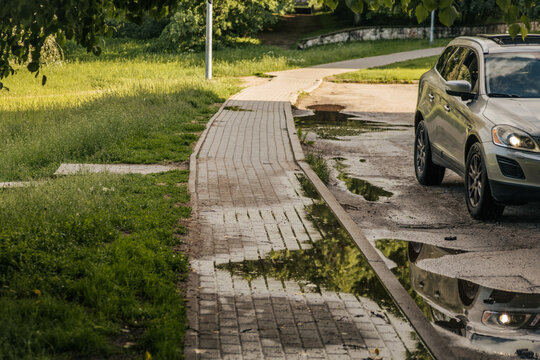 Silver car parked by a wet sidewalk with puddles, grass, and leafy trees in warm sunlight.