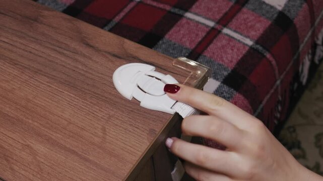 Woman's hand with red nail polish opens and closes a white child safety strap lock. Close-up shot shows the mechanism of the baby proofing furniture latch.