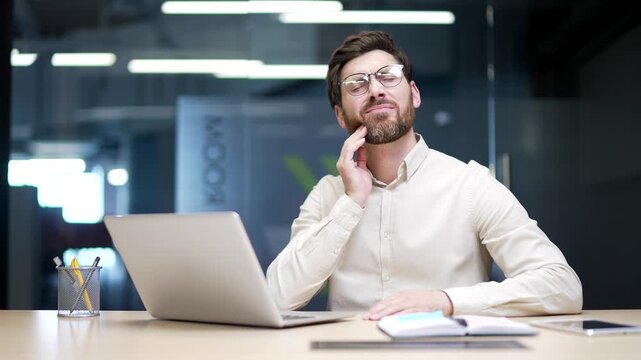 Businessman sitting at desk in modern office suffering from toothache. Male employee holding hand to cheek, feeling pain and discomfort while trying to work, suffering from dental or nerve problem.